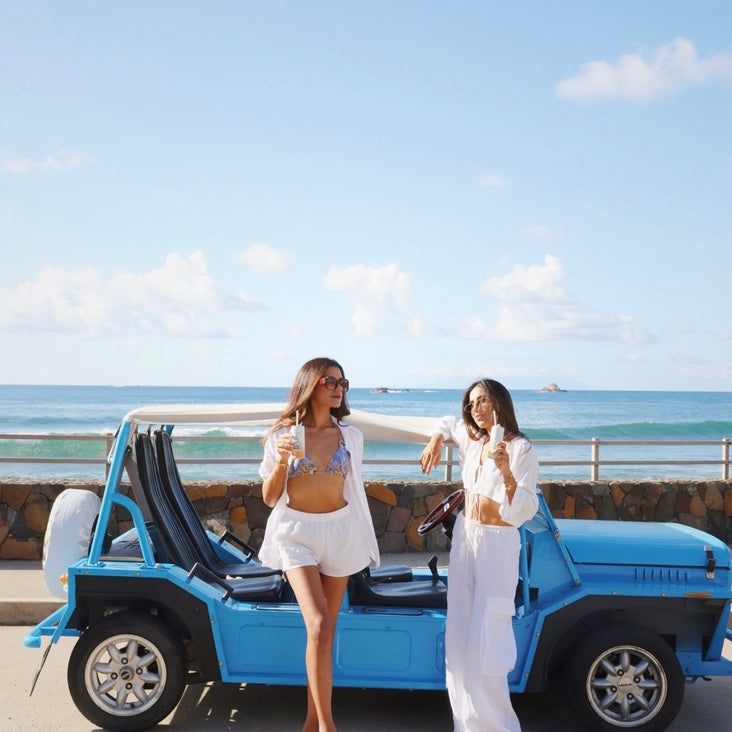 Two women standing next to a blue vehicle by the ocean.