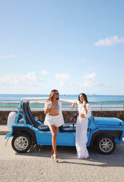 Two women standing next to a blue vehicle by the ocean.